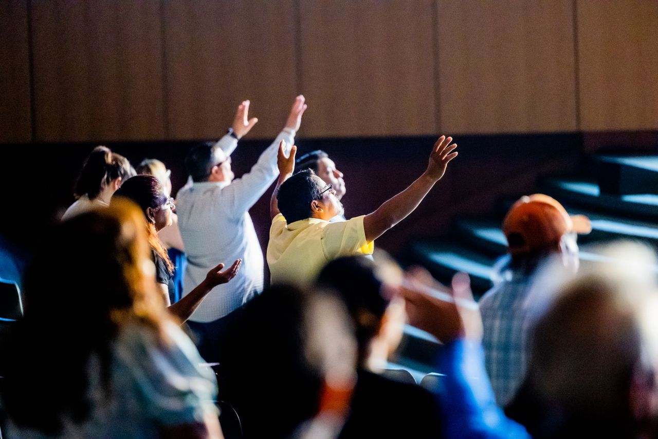 Home A diverse crowd engaged in worship indoors, raising hands in collective faith and devotion.
